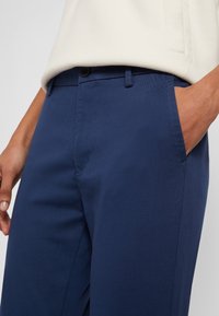 Granatowe spodnie z bawełnianej mieszanki, charakteryzujące się gładką fakturą, przednimi kieszeniami i zapięciem na guzik. Zbliżenie na bok i pas.