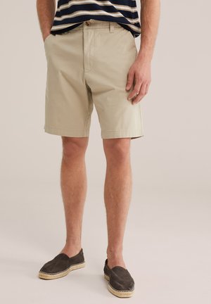 Mees, kes kannab beeže lühikesi pükse, triibulist särki ja tumedaid slip-on espadrille, seisab ühevärvilise tausta ees.