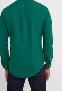 Camisa verde de manga larga con pinza en la espalda, puños abotonados, dobladillo curvado y tela de algodón suave. Sin patrones ni gráficos visibles.