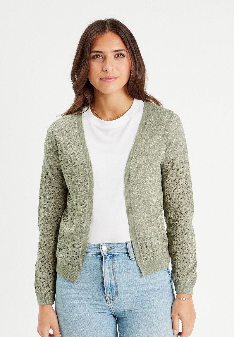 Hellgrüner Strickcardigan mit einem lockeren, offenen Strickdesign, kontrastierendem, einfarbigen Besatz und langen Ärmeln, über einem weißen Shirt getragen.