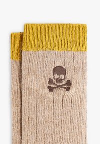 Guantes de lana beige con puños amarillos acanalados, que presentan un diseño de calavera y tibias cruzadas en marrón en la parte posterior de la mano.