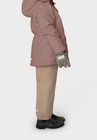 Veste isolante rose avec capuche, pantalon beige et gants gris. Matériaux texturés, poignets élastiques et chaussures noires. Vue de côté.