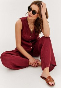 Burgundowa jumpsuit z lnu z guzikami z przodu, szerokimi nogawkami i bocznymi kieszeniami. Noszona z brązowymi klapkami i czarnymi okularami słonecznymi.