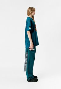 Camiseta de gran tamaño en color aqua con diseño bordado, combinada con pantalones a juego que cuentan con un bolsillo lateral y una etiqueta decorativa. Zapatos de goma negros.