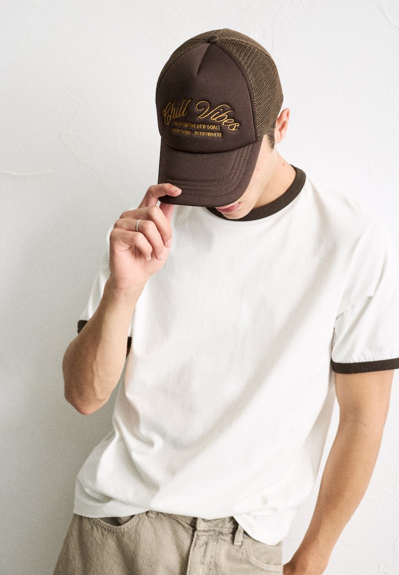 Brown mesh-back cap with embroidered "Chill Vibes" in gold, featuring a rounded front and contrast stitching, worn with a white T-shirt.