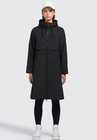 Long manteau noir à capuche avec fermeture éclair, taille élastique et deux poches latérales, associé à des leggings noirs et des baskets blanches.