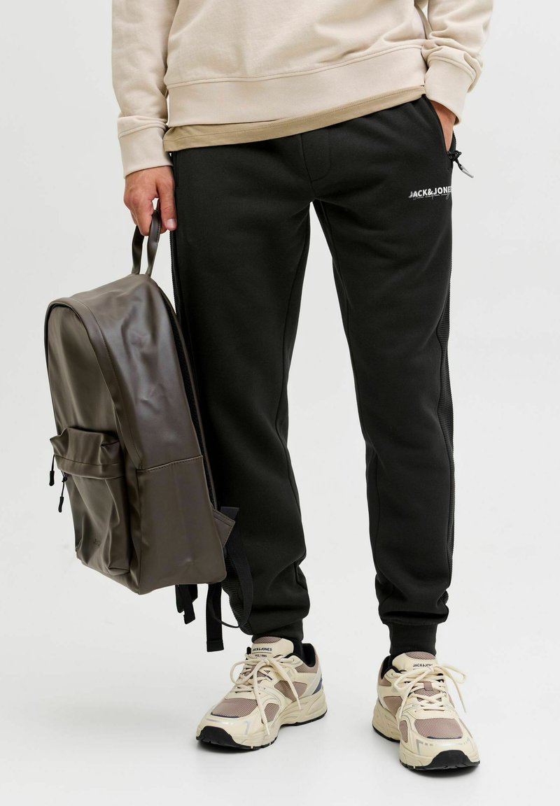Zwarte trainingsbroek met elastische tailleband, een beige sweatshirt en een donkerbruine rugzak. Crème kleurige sneakers met gaas en textuuraccenten.