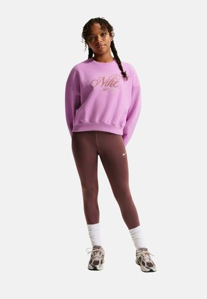 Joven mujer con cabello trenzado lleva una sudadera rosa de Nike, mallas marrones, calcetines blancos arrugados y zapatillas deportivas beige, está de pie relajada.