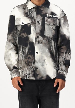 Mężczyzna noszący kurtkę z motywem tie-dye w kolorach czarnym, białym i szarym, z dwoma przednimi kieszeniami z klapką oraz logo "Carlo Colucci" na piersi.