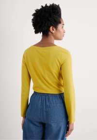 Kollane pika varrukaga topp avara kaelusega, kaasas vabade siniste teksapükstega, millel on elastne vöökoht ja küljetaskud.