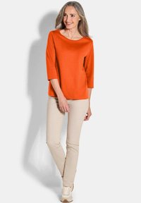 Oranger Pullover mit Dreiviertelärmeln, kombiniert mit beigen Hosen und hellen Sneakern. Glatte Textur, schlichtes Design und bequeme Passform.