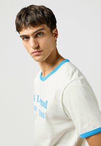 Camiseta blanca de manga corta, con cuello redondo acanalado azul y texto bordado en azul claro. Material de algodón suave con una textura lisa.