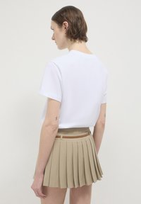 Frau mit kurzen braunen Haaren, die ein weißes T-Shirt und einen beigen plissierten Rock mit einem dünnen braunen Gürtel trägt, mit dem Rücken zu einem schlichten Hintergrund gewandt.