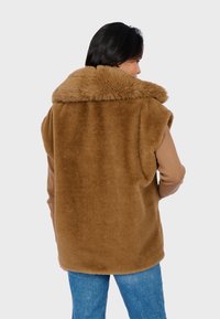 Gilet in finta pelliccia marrone con un ampio colletto strutturato, design senza maniche e un interno liscio, abbinato a jeans in denim azzurri.