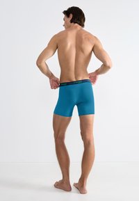 Bokserki w kolorze morskim, wykonane z gładkiego materiału, z czarnym pasem w talii z zielonym napisem logo, noszone przez modela od tyłu.