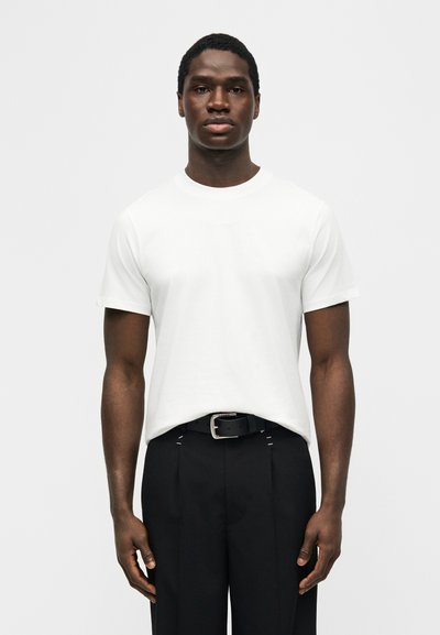 Jeune homme debout, face à nous, portant un t-shirt blanc uni à manches courtes rentré dans un pantalon noir avec une ceinture noire.