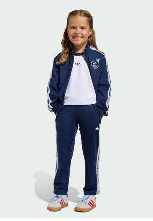 Navy tracksuit with white side stripes, featuring a heart logo on the jacket. White t-shirt underneath, paired with sneakers with red stripes.