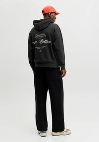 Mustast puuvillast kapuutsiga dressipluus tagaküljel graafilise tekstiga, kombineeritud lahtiste mustade pükste ja beežide tossudega. Modellil on peas punane nokamüts.