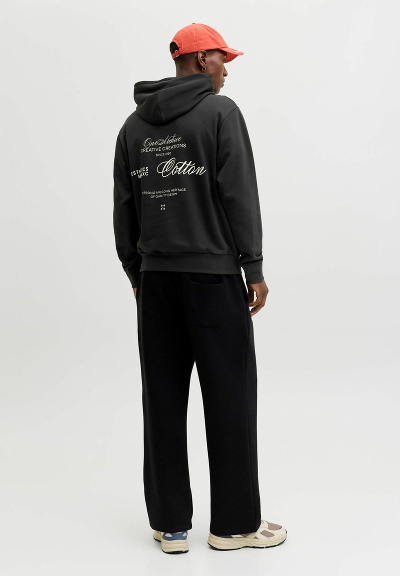 Mustast puuvillast kapuutsiga dressipluus tagaküljel graafilise tekstiga, kombineeritud lahtiste mustade pükste ja beežide tossudega. Modellil on peas punane nokamüts.