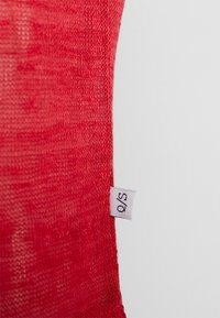 Tessuto a maglia rossa con una texture liscia. Presenta un piccolo etichetta bianca con la lettera "S" attaccata al bordo.