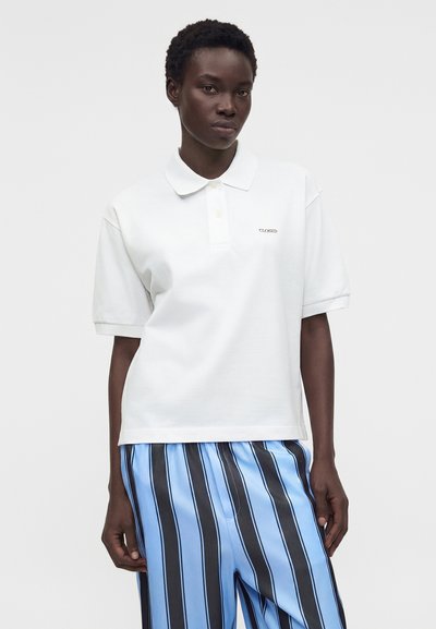 Γυναίκα με κοντά μαλλιά που φοράει λευκό πόλο με γιακά και μπλε παντελόνι με μαύρες και λευκές κάθετες ρίγες σε απλό φόντο.