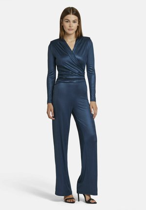 Marineblauer Jumpsuit mit langen Ärmeln, Wickeldesign und weit geschnittenem Bein. Aus einem glänzenden, glatten Stoff gefertigt. Getragen mit schwarzen High Heels.