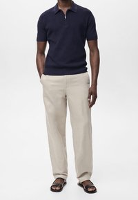 Polo bleu marine à manches courtes avec col zippé, associé à un pantalon large beige clair et des sandales noires. Texture lisse, coupe décontractée.