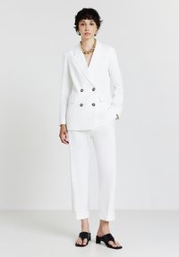 Donna con capelli corti e ricci, indossa un blazer bianco doppiopetto e pantaloni, una collana di perline dorate e sandali neri con tacco aperto.