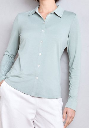 Person wearing a light green long-sleeve buttoned shirt with collar and white pants, standing against a plain light background.