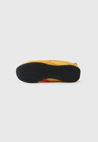 Sárga sneaker piros és szürke részletekkel. Gumi talp, texturált tapadással és sima felső anyaggal. Alacsony profilú kialakítás, lekerekített orral.