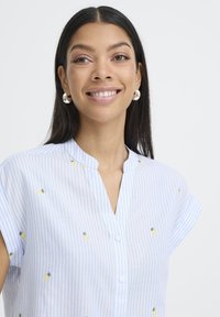Femme souriante aux longs cheveux foncés, portant des boucles d’oreilles anneaux argentées et une chemise à rayures bleu clair avec de petits motifs ananas.