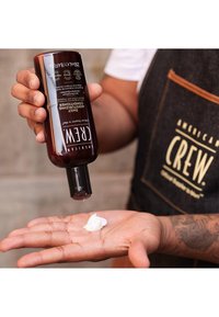 Brun flaske med svart hette som dispenserer hvit krem på en hånd. Produktetiketten har "American Crew" i fet skrift med instruksjoner og volumdetaljer.