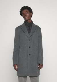 Cappotto grigio in lana con rever a punta, quattro bottoni neri e tasche laterali. Indossato sopra un giubbotto imbottito nero, con colletto zip.