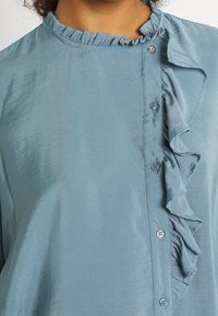 Light blue blouse made of smooth fabric; features ruffled collar and placket, with button detailing along the front.