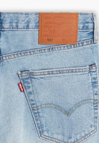 Vaqueros de mezclilla azul claro con una parche de cuero marrón con texto en relieve. Incluyen una etiqueta de logo roja en el bolsillo trasero y costuras en contraste.