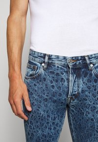 Jeans dżinsowe w jasnoniebieskim odcieniu, z wzorem w cętki lamparta, pięcioma kieszeniami, metalowymi elementami oraz pomarańczowymi akcentami przyszycia.