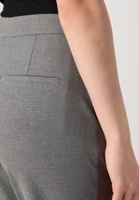 Gros plan d'une personne portant un pantalon gris sur mesure avec une poche arrière passepoilée et un haut noir, montrant le côté et le bras sur un fond neutre.