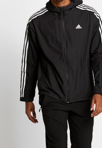 Svart lättviktsjacka med dragkedja fram, huva och vitt tre-stripes mönster på ärmarna. Har sidofickor och åtsittande manchetter.