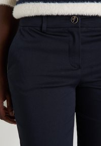 Pantalon bleu marine à la texture lisse, avec un bouton doré et une coupe ajustée. Comprend des poches avant et une fermeture éclair.