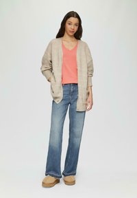 Beżowy sweter dzianinowy z kieszeniami noszony na koralowej koszulce z dekoltem w serek, w zestawieniu z niebieskimi dzwonami i beżowymi butami wsuwanymi. Widoczne strukturalne materiały.