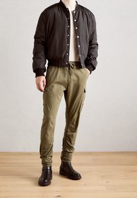 Schwarze Bomberjacke, cremefarbenes T-Shirt, olivfarbene Cargo-Hose mit elastischen Bündchen und braune Stiefeletten auf Holzboden.