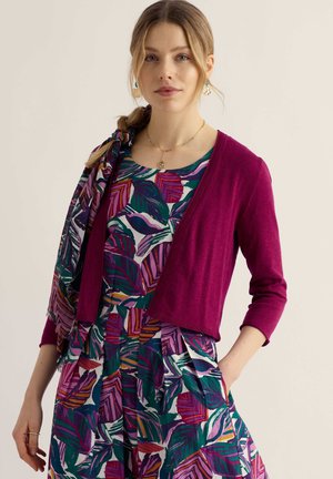 Young woman wearing a multicolored leaf-patterned dress with a magenta cardigan and matching scarf tied in her braided hair.