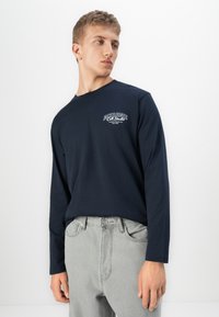 Ung man som bär en marinblå långärmad skjorta med vit text på bröstet och ljusgrå jeans, poserande framför en enkel vit bakgrund.
