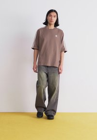 Tricou maro oversize cu un design alb în trei dungi pe mâneci, asortat cu blugi verzi lăsați și decolorati și încălțăminte neagră. Fundal simplu.