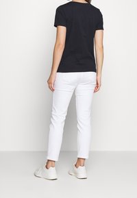 Žena v černém tričku s krátkým rukávem, bílých slim fit kalhotách a bílých teniskách, stojící proti jednoduchému světlému pozadí.