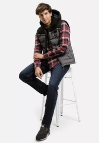 Kurtka puchowa w kolorze czarnym z kapturem, noszona na czerwono-czarną koszulę w kratę. Ciemne dżinsy i szare sneakersy dopełniają strój.