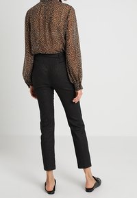 Kobieca bluzka w zwierzęcy wzór w odcieniach brązu i czerni z przezroczystego materiału, z buforowanymi rękawami, w zestawie z teksturowanymi czarnymi spodniami oraz płaskimi czarnymi butami.