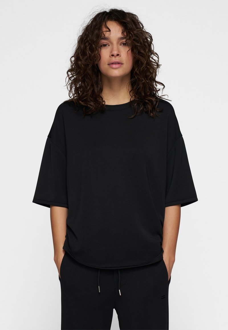 T-shirt nera oversize con maniche corte, scollo rotondo, texture liscia e vestibilità rilassata, indossata con pantaloni neri abbinati.