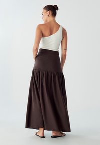 Hnedá maxi sukňa s perírovým dizajnom a drapovanou textúrou, v kombinácii s priliehavým bielym topom na jedno rameno. Modelka je zobrazená zozadu.