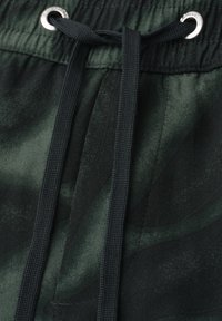 Dunkelgrüner Camouflage-Stoff mit schwarzen Kordeln, die durch silberne Metallösen am Bund der Hose gezogen sind.
